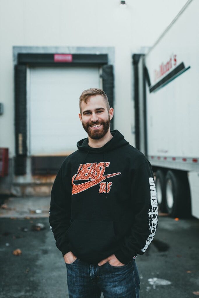 A smiling bearded man wearing a hoodie stands confidently outdoors by a truck.
